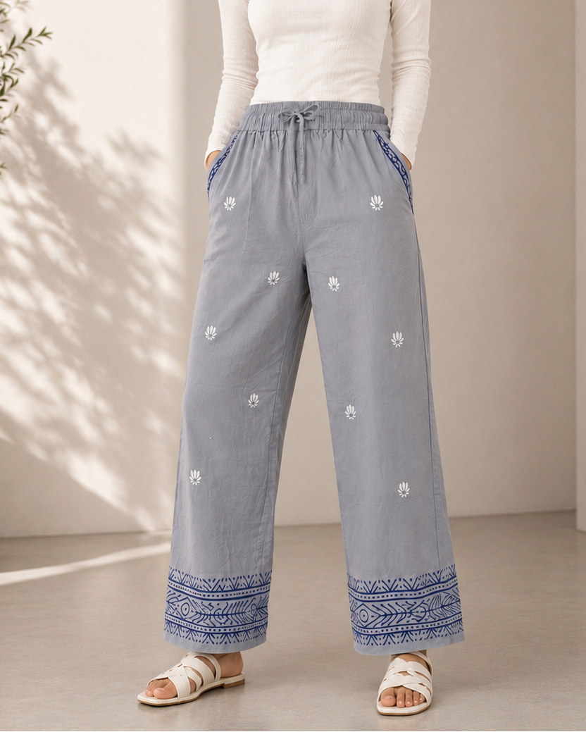 A woman modeling wide-leg light gray pants with blue patterned hems and white embroidered details, paired with a white long-sleeve top and white slide sandals in a sunlit indoor setting.