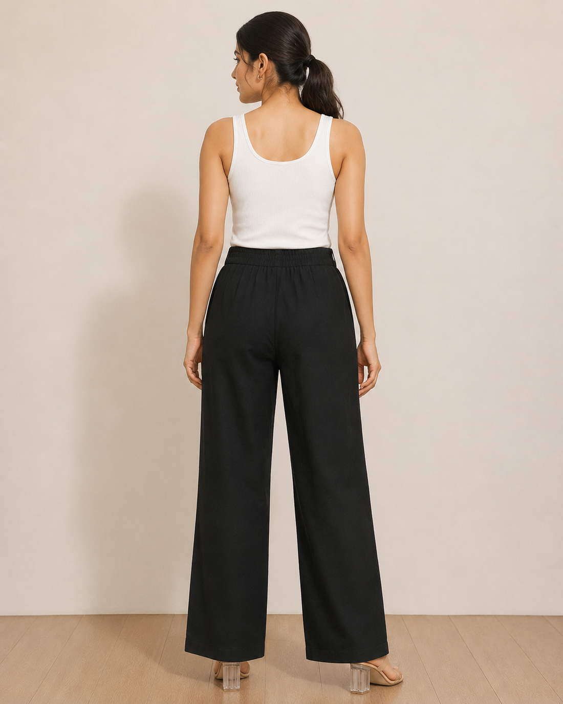 A woman modeling black wide-leg pants with white embroidered tree and bird motifs, paired with a white ribbed tank top, standing against a neutral beige wall.