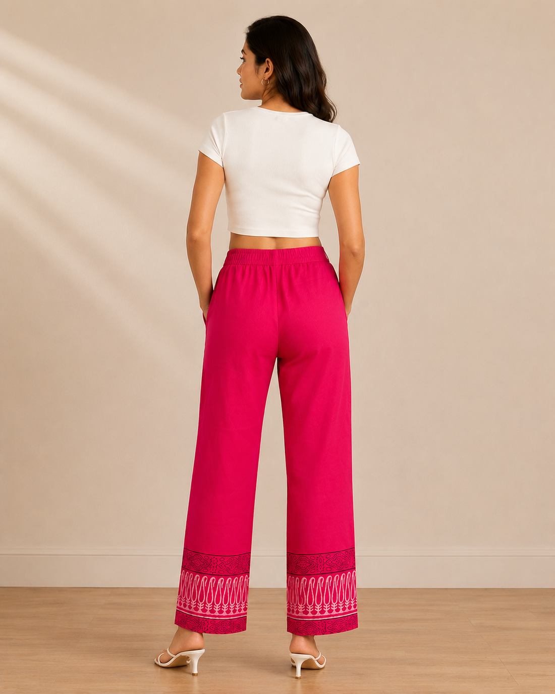 A woman modeling bright pink wide-leg pants with white printed motifs and a patterned hem, paired with a white crop top and clear high-heel sandals, against a beige studio backdrop.