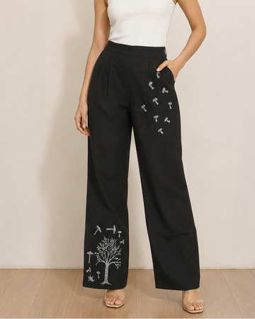 A woman modeling black wide-leg pants with white embroidered tree and bird motifs, paired with a white ribbed tank top, standing against a neutral beige wall.