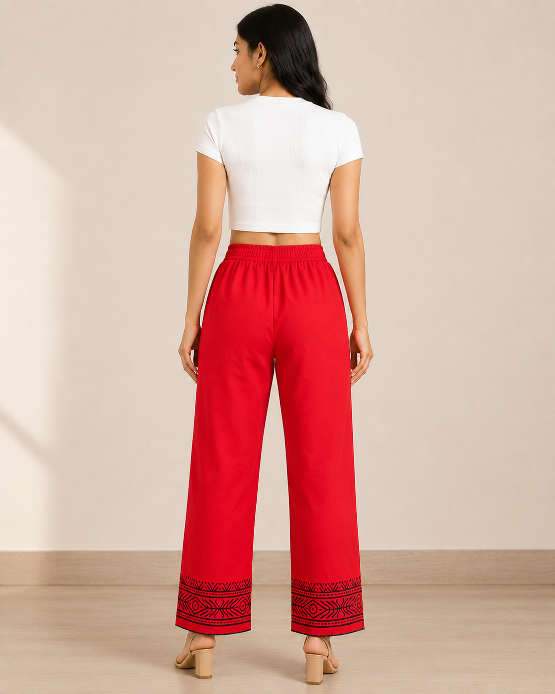 A woman modeling red wide-leg cropped pants with black embroidered floral details and a black patterned hem, paired with a white crop top against a beige studio background.