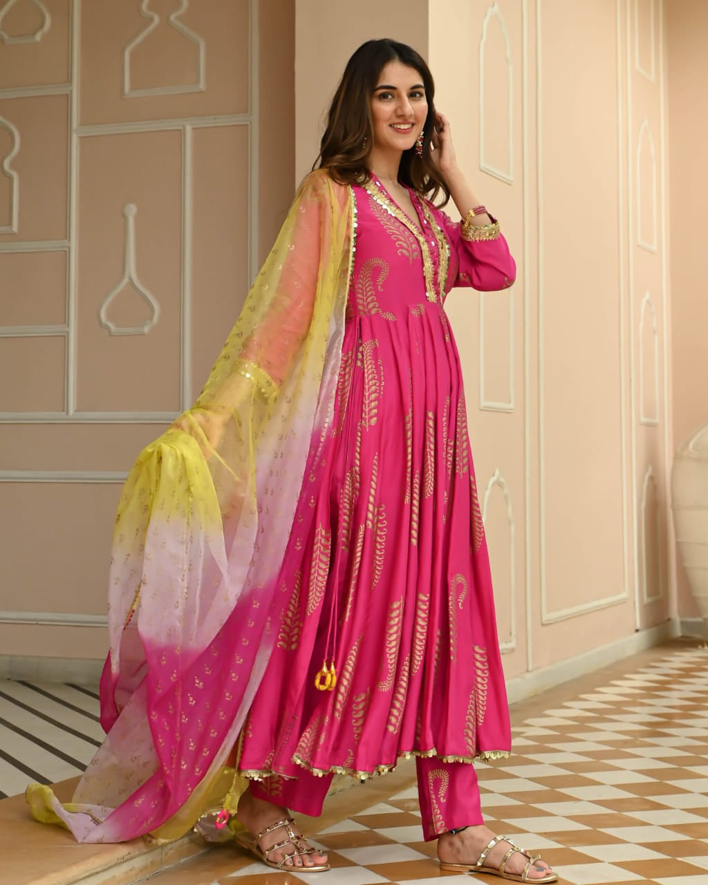 A woman models a bright pink traditional Anarkali suit with gold embroidery, matching pants, and a sheer yellow-to-pink gradient dupatta, set against a beige wall with decorative arches and a checkered floor.