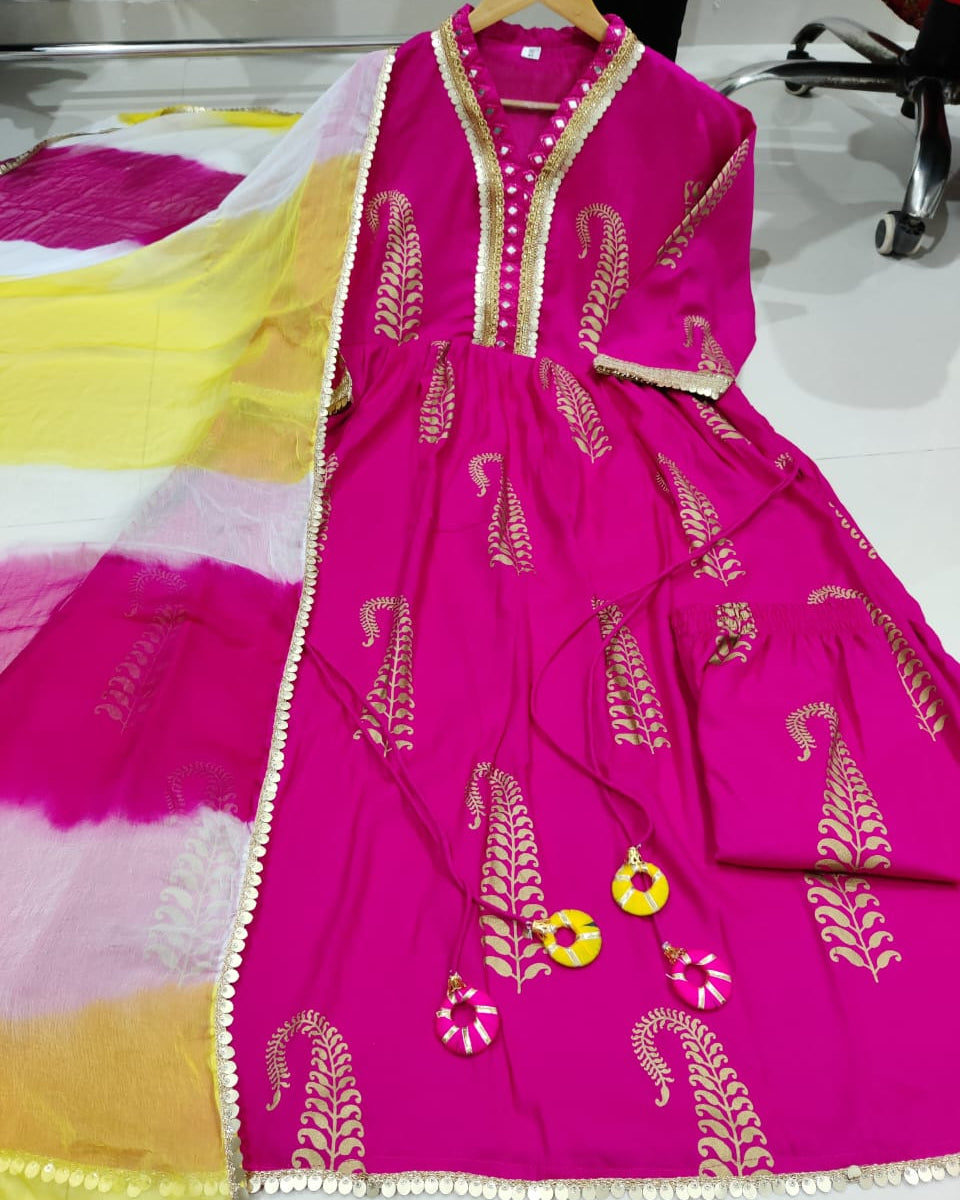 A bright pink traditional Anarkali gown with gold leaf prints and a matching sheer yellow and pink dupatta, displayed on a hanger on a white floor.