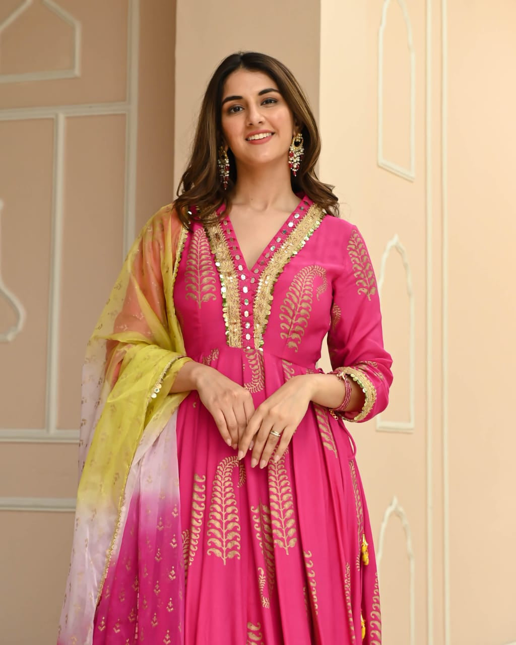 Woman wearing a pink embroidered dress with a yellow dupatta against a beige wall.