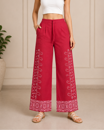 A woman modeling high-waisted, wide-leg trousers in a vibrant magenta pink with white embroidered paisley patterns down the sides and at the hem, paired with a white crop top and beige flat sandals.