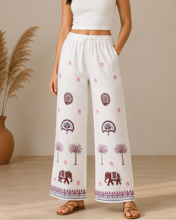 A woman modeling white wide-leg pants with pink and brown bohemian-style prints, paired with a white crop top, standing in a studio with a beige background and a pampas grass vase.