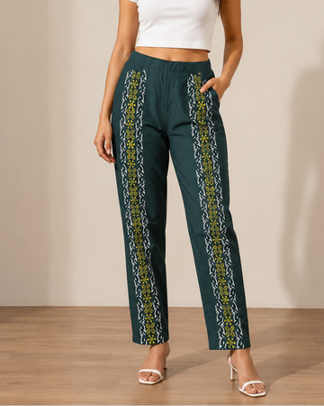 A woman modeling a pair of dark teal wide-leg trousers with vertical yellow and white patterned side panels, paired with a white crop top and white heeled sandals, standing against a beige studio wall.