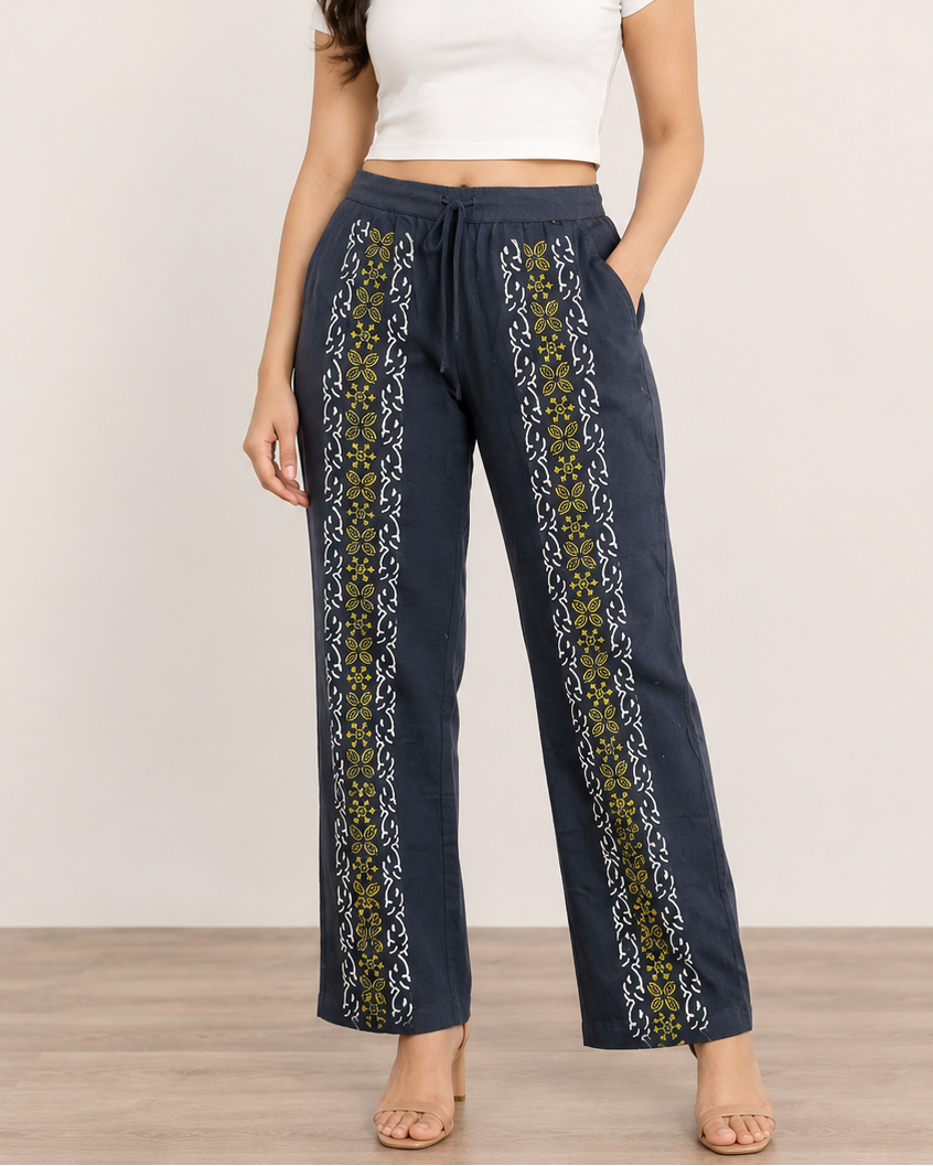 A woman modeling a pair of dark navy wide-leg pants with vertical yellow and white patterned side panels, paired with a white crop top against a neutral studio background.
