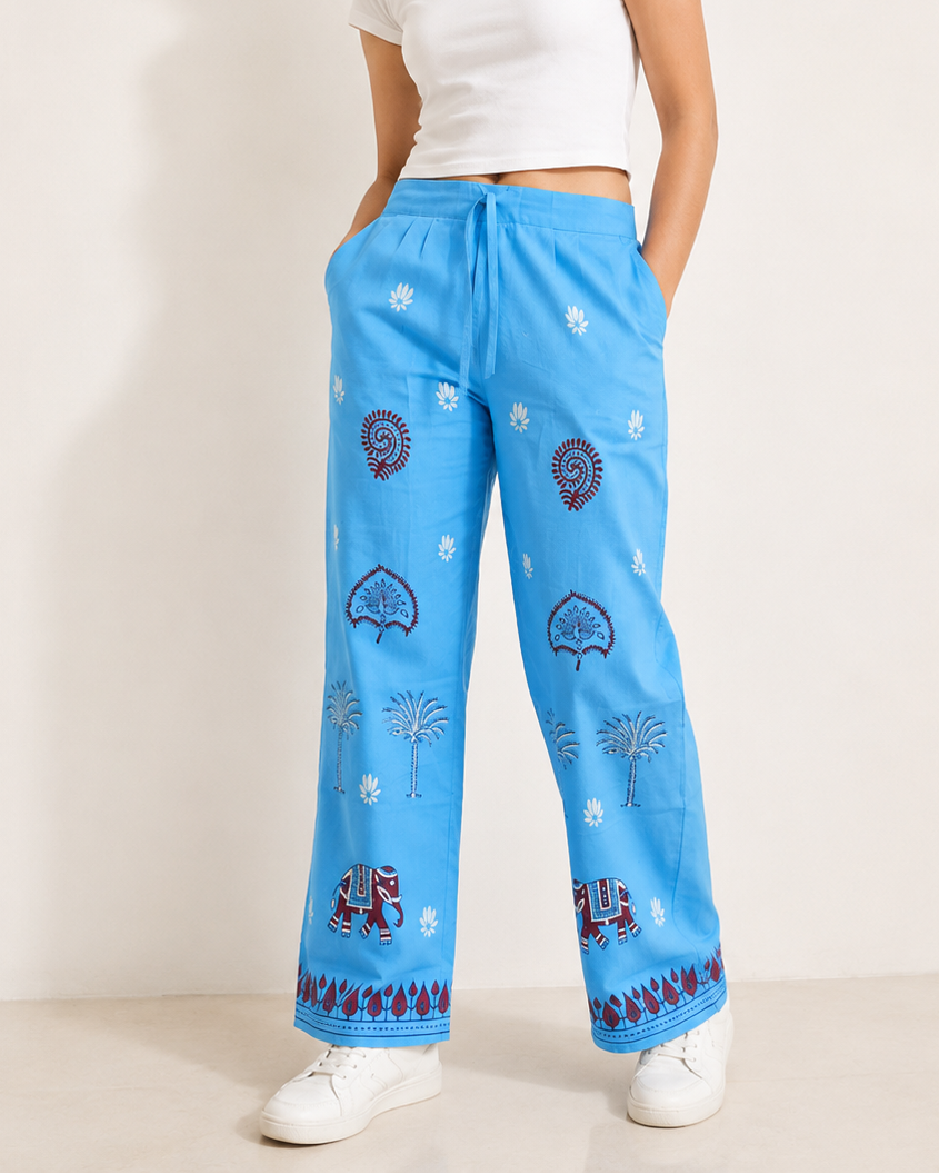 A woman modeling bright blue wide-leg pajama pants with a white crop top and white sneakers against a plain beige studio background.