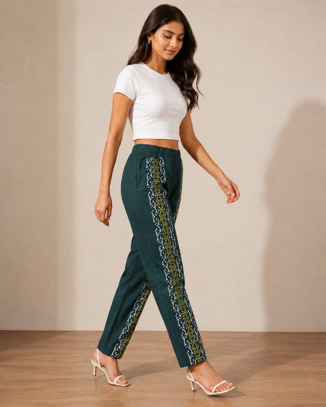 A woman modeling a pair of dark teal wide-leg trousers with vertical yellow and white patterned side panels, paired with a white crop top and white heeled sandals, standing against a beige studio wall.